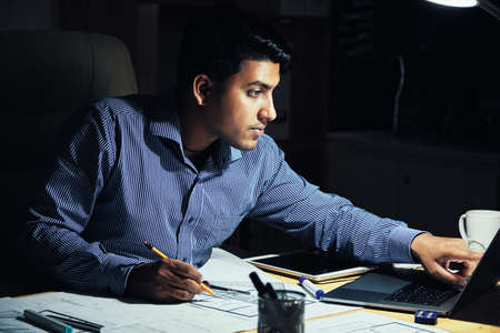 Young Indian civil engineer checking data on laptop when drawing plan of apartmentの写真素材