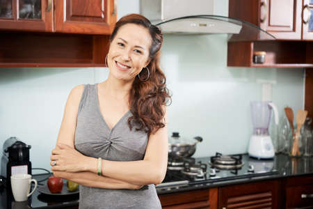 Cheerful middle-aged woman standing in kitchen with her arms foldedの写真素材