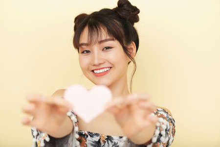 Wonderful smiling Asian woman with hair buns outstretching hands with paper heart on yellow backgroundの写真素材
