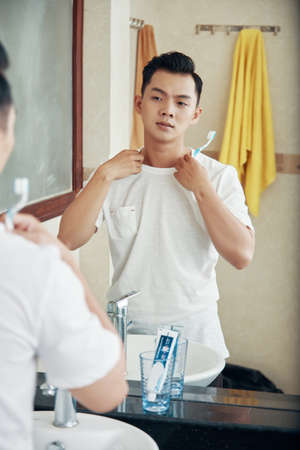 Young modern Asian man holding toothbrush and looking at himself in reflection of bathroom mirrorの写真素材