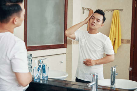 Young Asian man grooming hairstyle in mirror reflection standing in bathroom at homeの写真素材