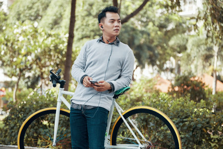 Attractive Asian guy holding smartphone and listening to music while leaning on bicycle on...