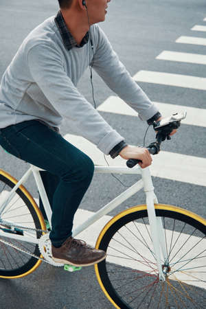 Crop guy in casual outfit listening to music in earphones and looking away while riding bicycle near crosswalk on city roadの写真素材