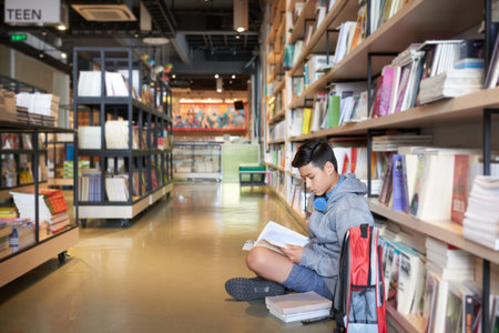 Side view of teenage boy with headphones sitting on library floor with backpack leaning on bookshelves readingの写真素材