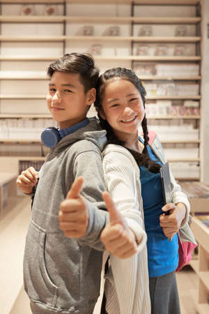 Portrait of happy Asian boy and girl standing back to back and giving thumbs up in school libraryの写真素材