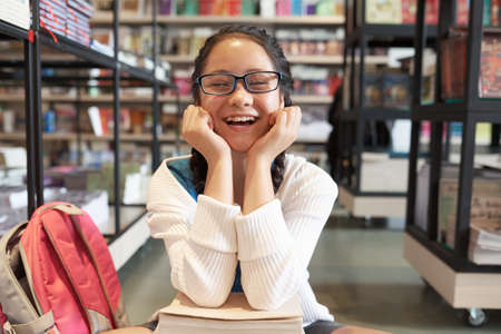 Girl in glasses sitting with head on hands smiling with closed eyes on blurred bookshelves backgroundの写真素材