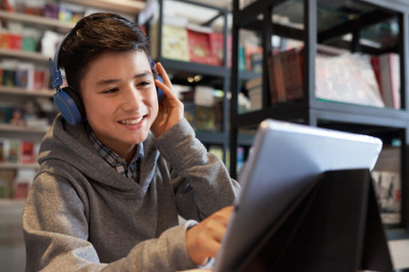 School boy sitting touching headphones looking at tablet smiling on bookshelves background