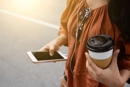 Hands of woman drinking take-out coffee and using music application on her smartphoneの写真素材