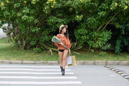 Young stylish woman with flowers at paper-bags crossing roadの写真素材