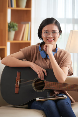 Portrait of cheerful Vietnamese girl in glasses playing guitar at homeの写真素材
