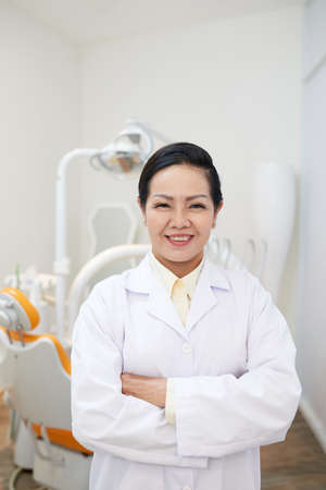 Adult Asian woman in white medical gown holding hands crossed and smiling at camera in dental officeの写真素材
