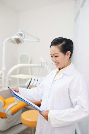 Professional modern Asian woman in white gown reading clipboard paper while standing in dental officeの写真素材