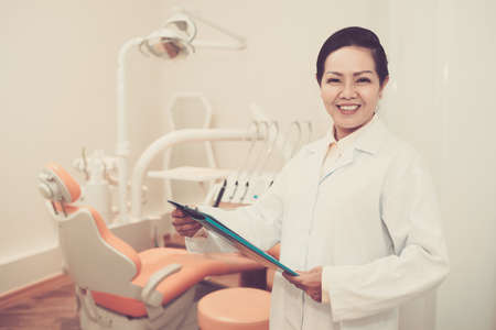 Cheerful Asian adult doctor in dental office holding clipboard and smiling at cameraの写真素材
