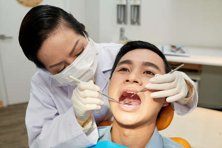Woman in mask and gloves exploring oral cavity of young Asian man in dentist officeの写真素材