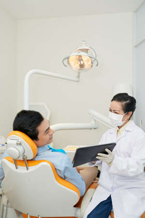 Adult dentist in mask showing tablet to young Asian man in dental chair during visit in officeの写真素材
