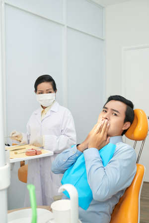 Adult Asian man covering mouth in worries looking at camera while sitting in dental chair with doctor nearの写真素材