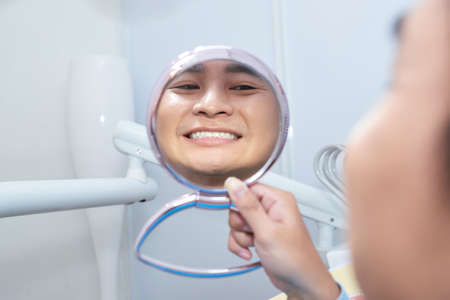 Adult Asian man checking white teeth in mirror reflection while sitting in dental chair in officeの写真素材