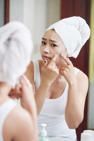 Young Asian woman in white towel on head popping pimple in mirror reflection standing in bathroomの写真素材