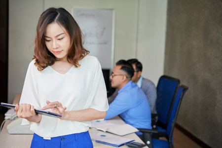 Beautiful Asian woman standing in office room with coworkers on background and using tablet