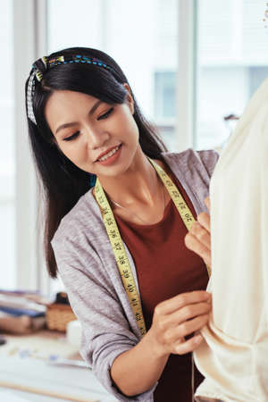 Pretty Asian female smiling and adjusting delicate blouse on mannequin while working inの写真素材
