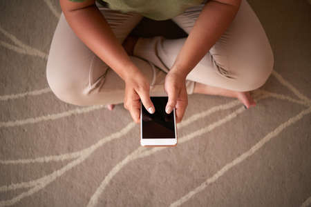 From above shot of anonymous female sitting on floor and browsing smartphone with blank screenの写真素材
