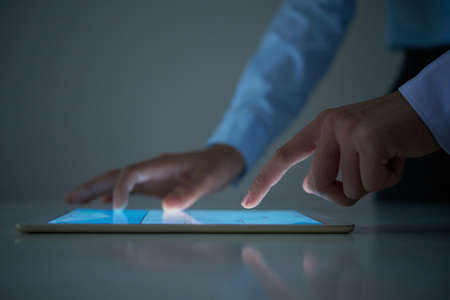 Crop hands of unrecognizable businessman enlarging and scrolling data on modern tablet on table in dark officeの写真素材