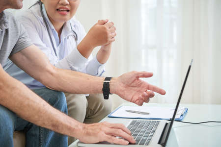 Crop shot of elderly man showing laptop to female doctor sitting at table at homeの写真素材