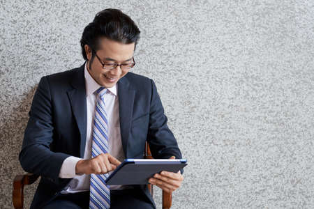 Young Asian man in suit and eyeglasses using tablet while sitting in armchair on gray wallの写真素材
