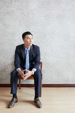 Adult Asian man in classy suit sitting on chair and looking away in thoughts on gray backgroundの写真素材
