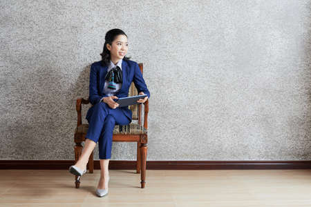 Beautiful modern businesswoman sitting in armchair with tablet looking awayの写真素材