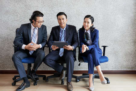 Elegant Asian woman and men sitting together on office chairs with tablet and looking at cameraの写真素材