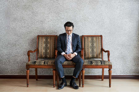 Serious focused Asian man in formal suit sitting on chair looking down in deep thoughtsの写真素材