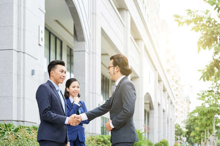 Side view of Asian businessmen shaking hands on street with young female colleague near in sunlightの写真素材
