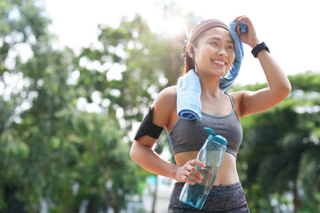 Young happy Asian woman in sportswear wiping forehead with towel while smiling away in park