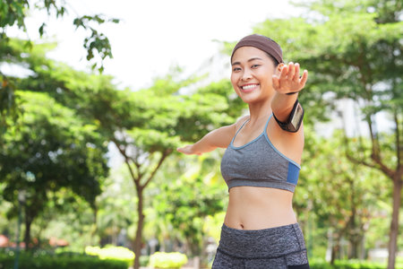 Young happy Asian woman in sportswear and armband holding hands apart and training in summer park