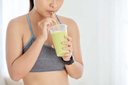 Crop shot of sportive woman drinking green smoothie with straw standing indoorsの写真素材
