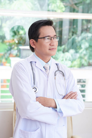 Confident Asian doctor standing in lab coat and eyeglasses at his officeの写真素材