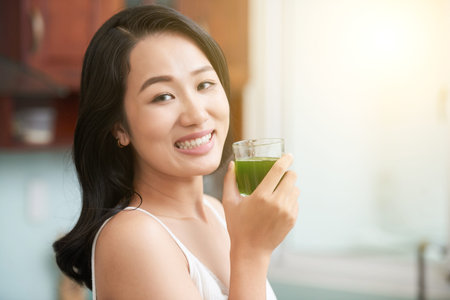 Smiling Asian woman holding glass with fresh green juice being healthy and smiling at cameraの写真素材