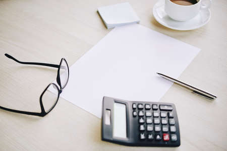 Office table with calculator, spectacle, a cup of coffee, a pen and a blank paperの写真素材