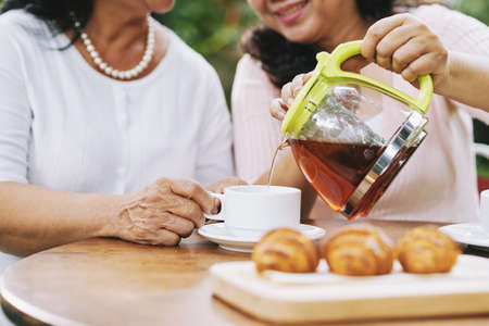 Close-up view of unrecognizable Asian women sitting at table with croissants, pouring fresh tea from pot, talking and smilingの写真素材