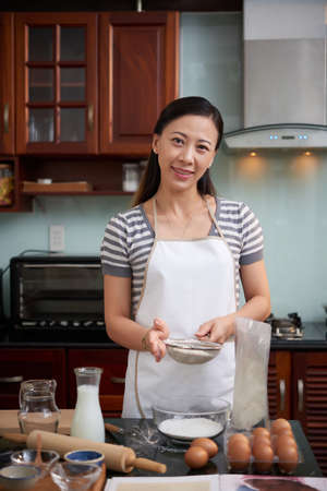 Portrait of young attractive Asian woman smiling at camera joyfully while standing at kitchen counter and sifting flour for cookie mixtureの写真素材