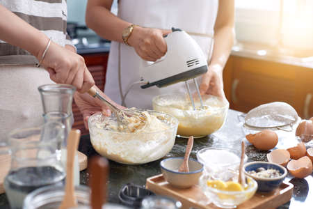 Unrecognizable women making cookie mixture in kitchen, using whisk and modern electric mixer while beating eggsの写真素材