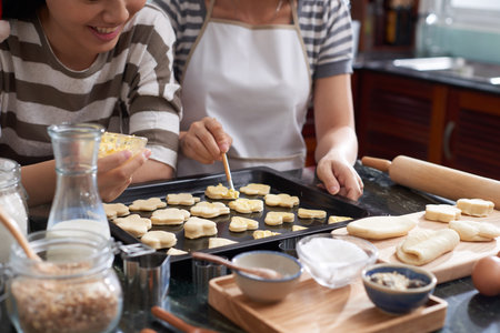 Crop shot of unrecognizable women standing by kitchen table and spreading butter on cut out raw...