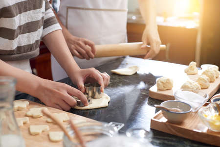 Unrecognizable woman rolling cookie mixture while another cutting out cookies with cutters in sunlit home kitchenの写真素材