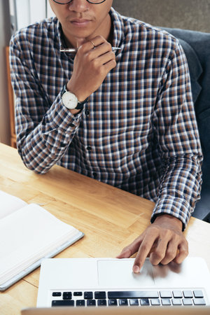 Unrecognizable young man in checked shirt sitting at table and reading information on notebook  with...
