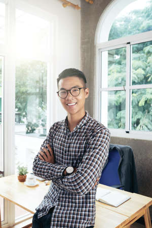 Portrait of young handsome Asian man standing in bright office with crossed hands, leaning on table and smiling at cameraの写真素材
