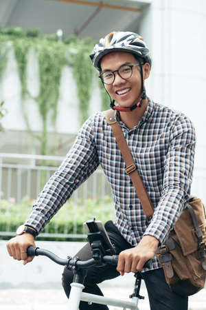 Portrait of handsome Asian guy wearing glasses and safety helmet sitting on bicycle outdoors and smiling at camera cheerfullyの写真素材