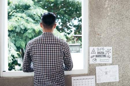 Rear view of unrecognizable dark-haired man looking out the window while  standing in office with...