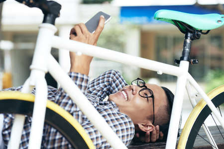 Young Asian man having video chat on smartphone and smiling cheerfully while lying outdoors behind his bicycleの写真素材