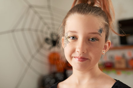 Portrait of beautiful little Caucasian girl with eye makeup and painted spider on her face smiling at cameraの写真素材